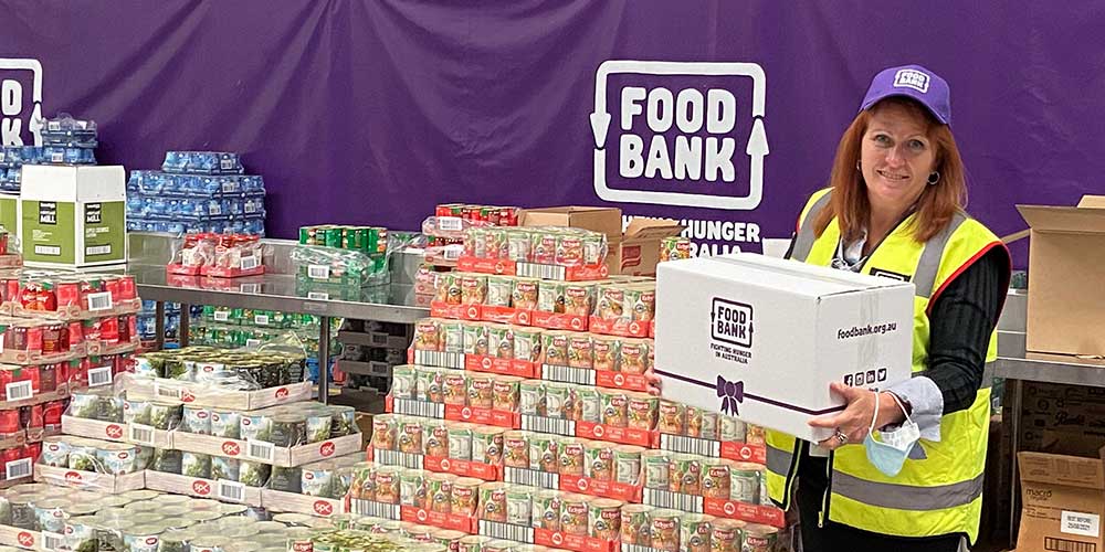 Kate O'Hara, CEO of Foodbank WA, holding a foodbank hamper in front of pallets of food products.