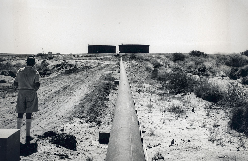 A black and white photo of a pipeline on Barrow Island running towards two silos.